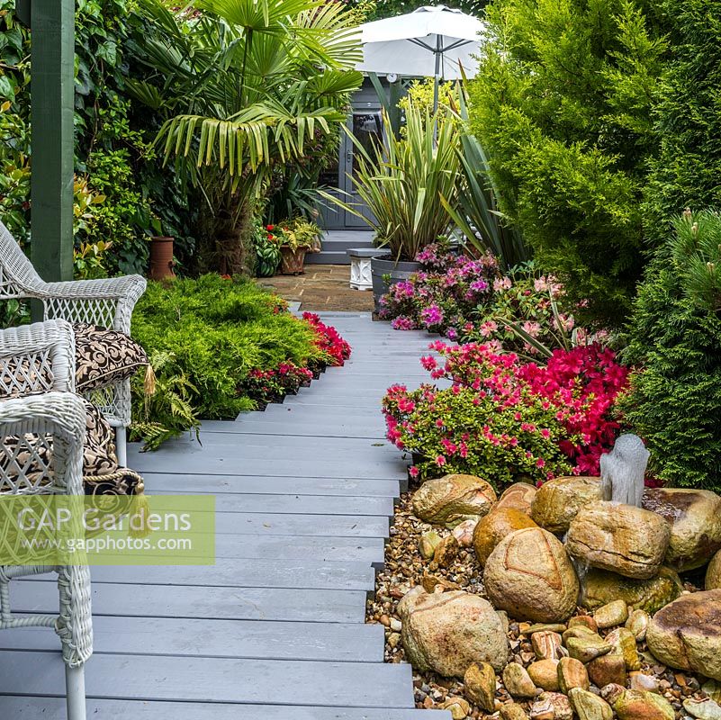 3.7m x 25m contemporary garden. Second, central seating area in shade of pergola,  linked to each end by stepped plank path. Evergreen planting includes flowering azaleas, phormium, golden cypress and Chusan palm.