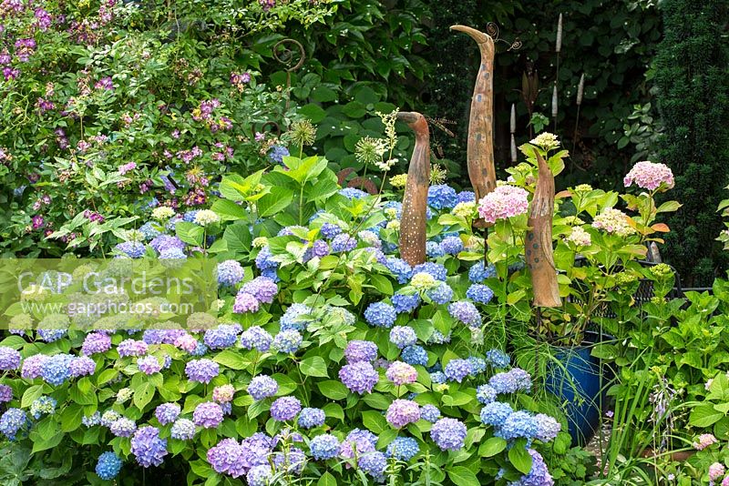 Three long shaped ceramic birds next to Rosa 'Veilchenblau' and Hydrangea macrophylla 'Endless Summer'