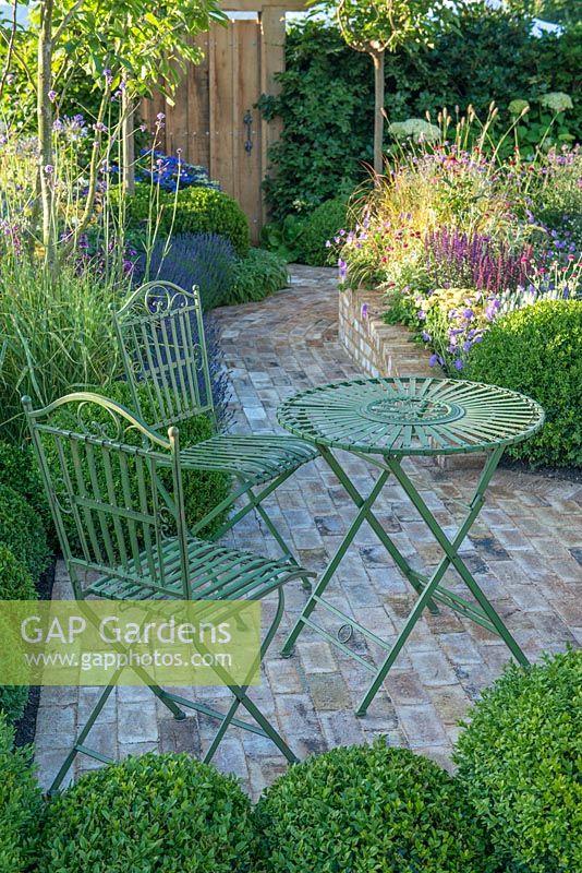 Wrought iron table and chairs on brick paving surrounded by box topiary balls - Squire's Garden Centres Urban Oasis - RHS Hampton Court Flower Show 2015