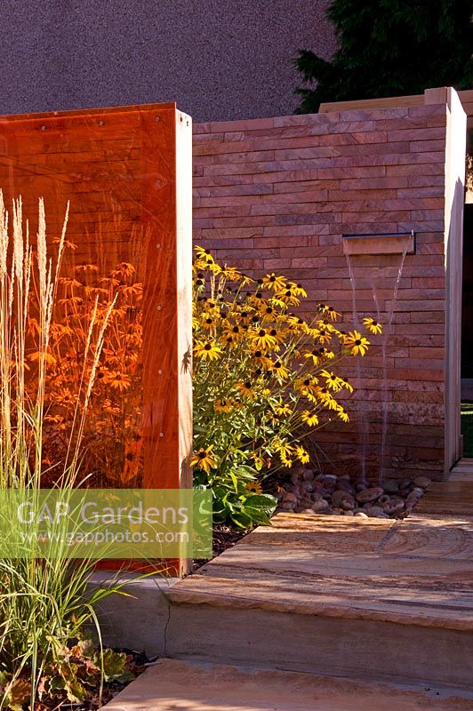 Small urban contemporary town garden with wooden deck path between border of grasses and Rudbeckia - orange perspex screen divider creating privacy wall with lip water feature waterfall cascade.  Ansari garden, Harrow