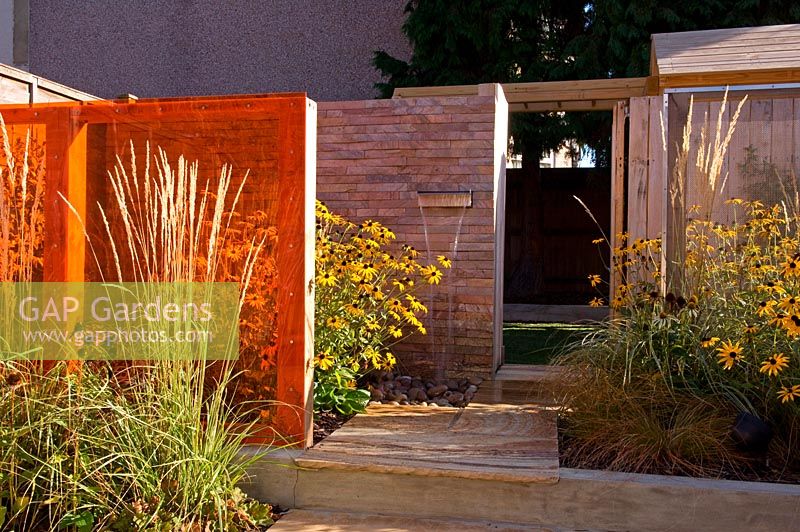 Small urban contemporary town garden with wooden deck path between border of grasses and Rudbeckia. An orange perspex screen divider creates privacy.  Brick wall with lip water feature - waterfall. Ansari garden, Harrow