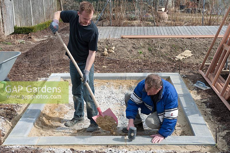 Men laying the concrete foundation for a greenhouse with shovel, sand and rubber mallet