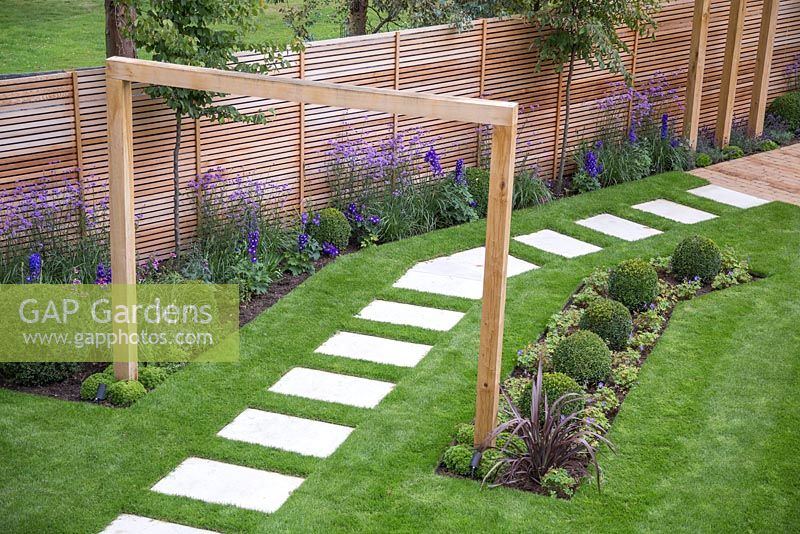 View of the path featuring white stone slabs, wooden beam entrance and borders with Verbena bonariensis, Buxus sempervirens, Delphinium elatum 'Blaustrahl' and Geranium 'Johnson's Blue'