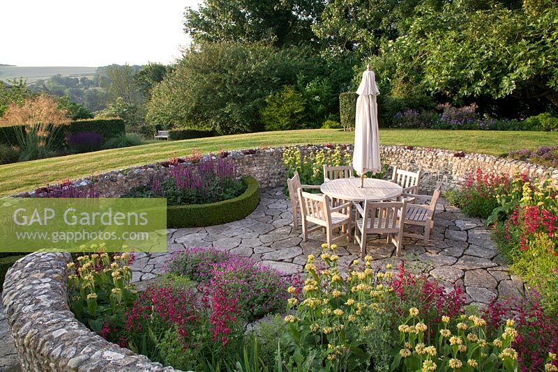 Table and chairs in sunken garden terrace with flint wall and planting of Penstemon, Phlomis, Salvia nemerosa 'Caradonna' and Geranium. Follers Manor, Sussex. Designed by: Ian Kitson