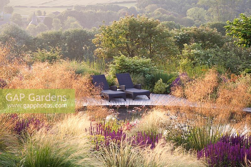 Sunloungers on wooden deck next to pond with Salvia, Stipa gigantea, Stipa tenuissima, Penstemon, Pontederia and Butomus umbellatus - flowering rush. Follers Manor, Sussex. Designed by: Ian Kitson