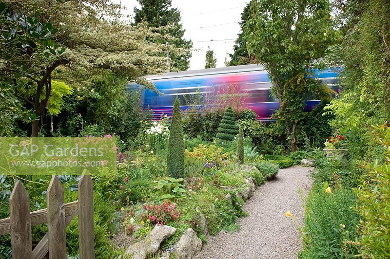 View from a garden gate down a gravel path beside rockery raised bed border with a train speeding by at the bottom of the garden 
