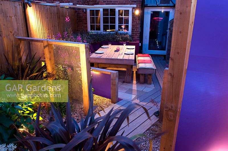 Contemporary long narrow small urban family garden with wooden decked paths at night. 