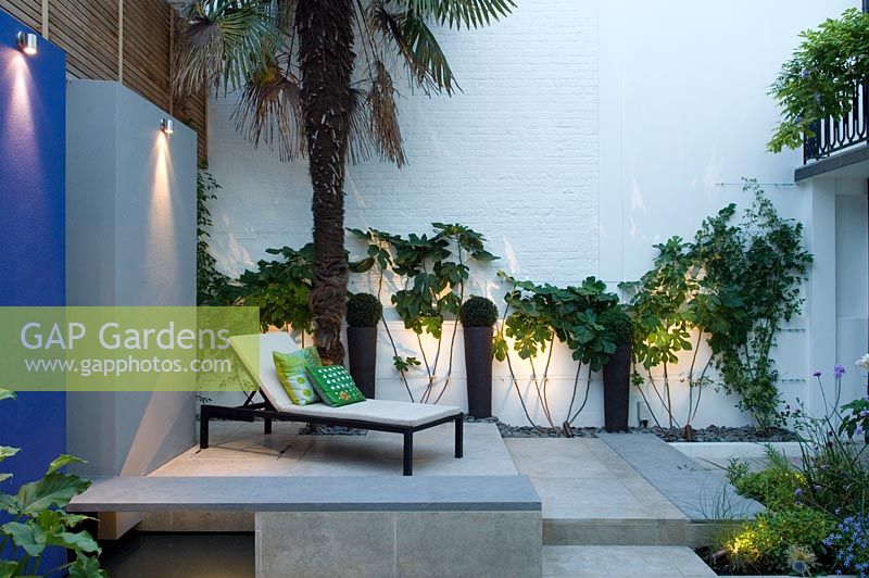 High walled small urban courtyard garden. Outdoor living room with painted wall panels, water feature and lounger on a raised level. 
