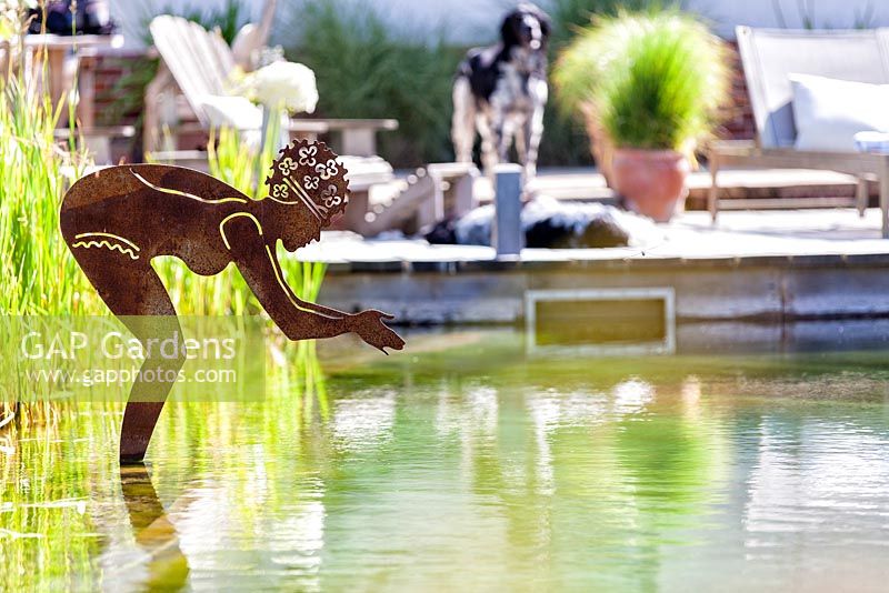 View of brushed steel diving lady sculpture with seating area and dogs in the background. 