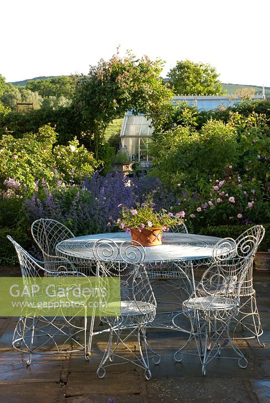 Metal table and chairs on a patio with view to greenhouse through Nepeta 'Six Hills Giant'