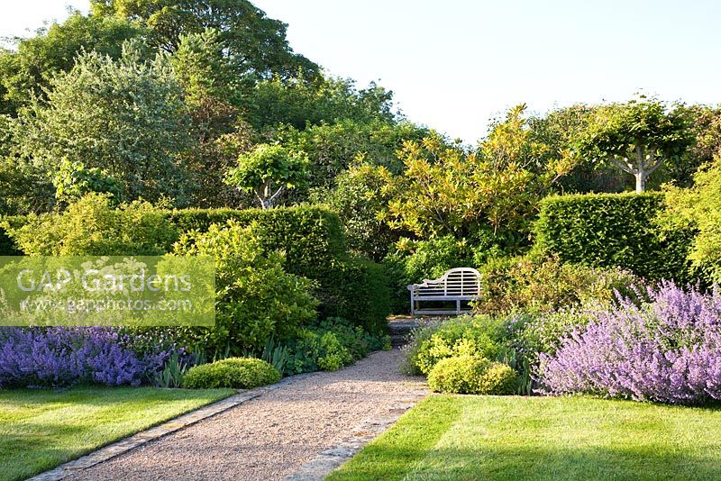 A wooden seat in a quiet corner of a garden with Roses, Nepeta, Geranium and Alchemilla mollis. Designer Georgia Langton. Farleigh House, Hampshire.