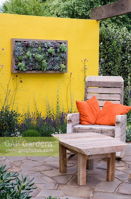 Vertical bed of Sempervivums, Euphorbia myrsinites and Sedums on a yellow painted wall, chair and table made from recycled pallets - Summer in the garden, RHS Hampton Court Flower Show 2012