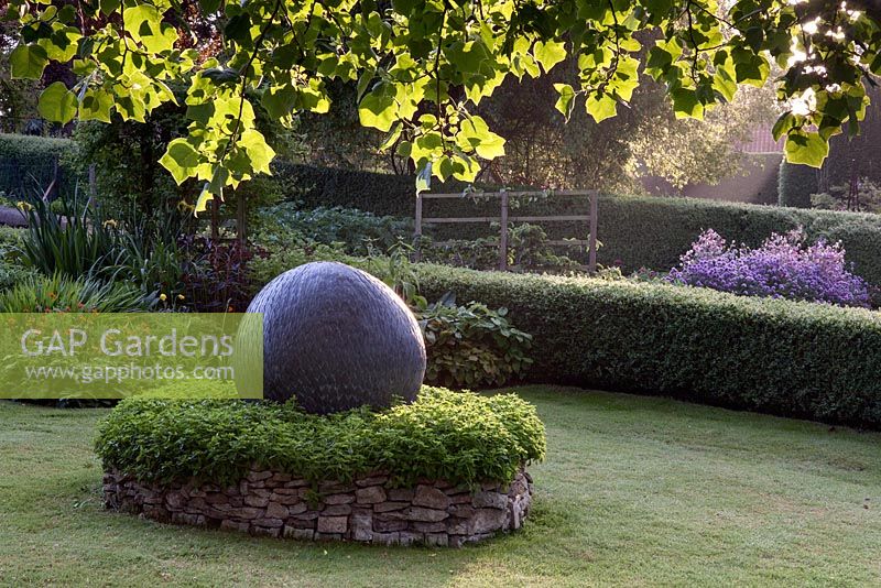 Under the Liriodendron tulipifera - Tulip Tree, looking towards the stone circle and zinc sphere, with part of the hot coloured herbaceous borders behind, and the vegetable garden beyond. Fawley House, North Cave, Yorkshire, UK. 
