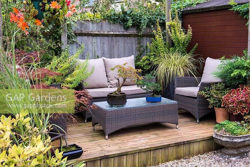 Tucked away in quiet corner at bottom of garden a raised wooden deck with outdoor rattan furniture and containers of acer, bonsai, verbena, coleus and ornamental grasses.