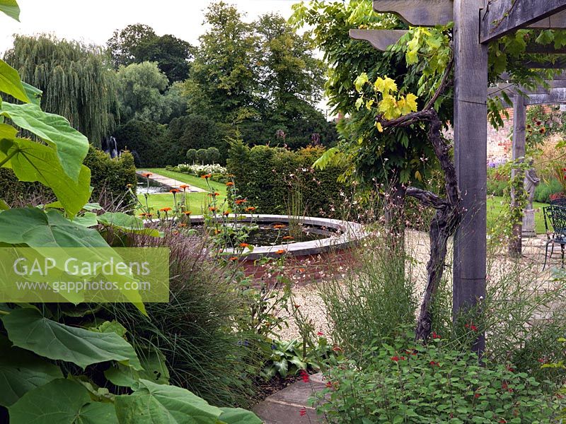 A view from the hot border with Wisteria, Vitis, Guara lindheimeri, Tithonia rotundifolia and Paulownia tomentosa to the raised brick pond and formal garden beyond.