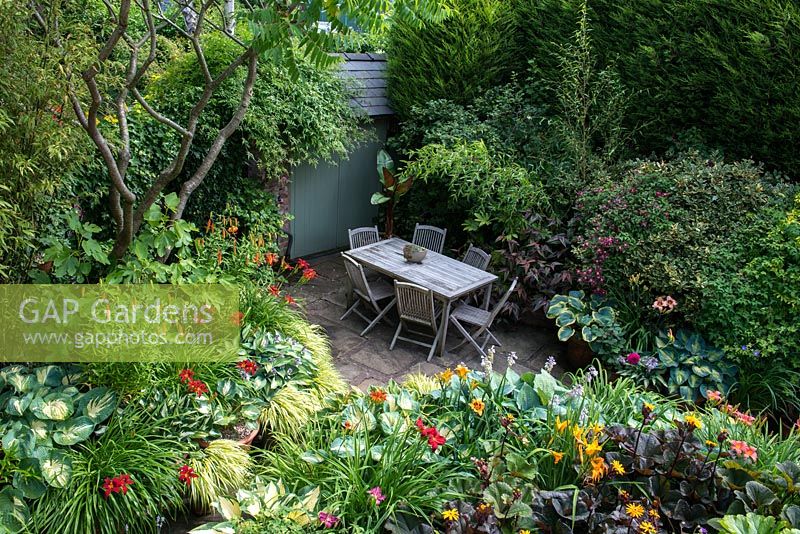 A secluded shady patio with garden furniture surrounded by lush dense borders planted with Hostas, Hakonechloa grasses, Ligularia, Persicaria microcephala, Lilium martagon, daylilies, Clematis Etoile Violette, Bamboo and a banana plant 