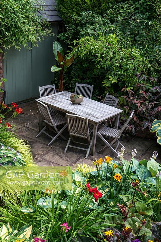 A secluded shady patio with garden furniture surrounded by lush dense borders planted with Hostas, Hakonechloa grasses, Ligularia, Persicaria microcephala, Lilium martagon, daylilies, Clematis Etoile Violette, Bamboo and a banana plant 
