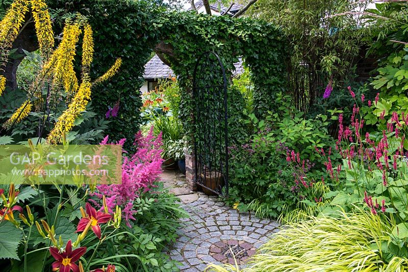 A ivy clad brick doorway leading to a secluded patio. In the foreground the borders are planted with Astilbe Fanal, Hemerocallis Crimson Pirate and Ligularia ligularia przewalskii, Astrantia, Hakonechloa macra Aureola and Persicaria 