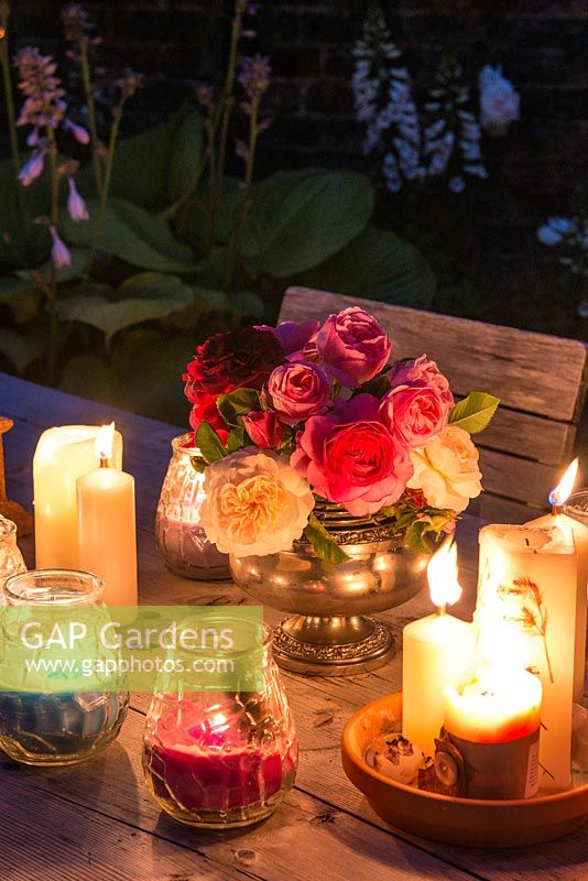 Table is prepared for summer barbecue with vase of roses and candles. Glass candleholders in borders illuminate planting  from left to right, Hosta 'Sum and Substance' and Digitalis alba.