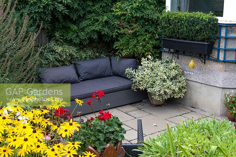 A sunken patio with outdoor sofa. Japanese painted ferns are planted in the shady area behing and containers with bright Pelargoniums providing long lasting colour.