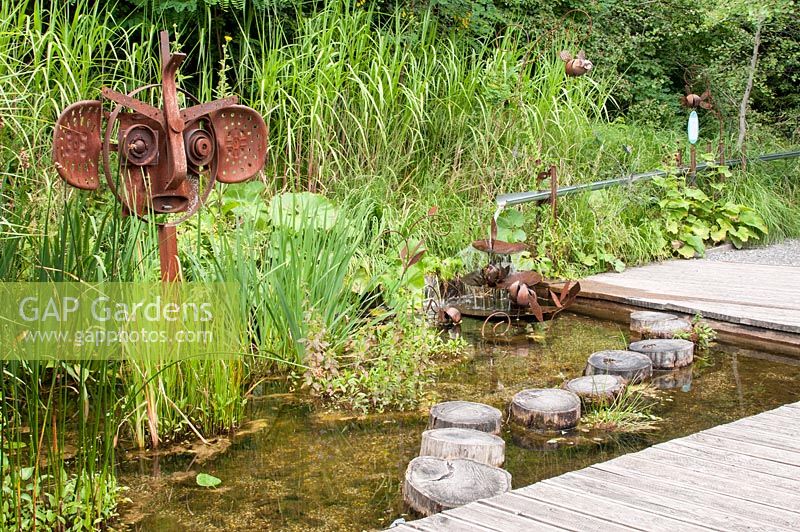 Natural clear shallow pond with water feature and sculpture made of rusted steel with adjacent decking stepping logs and water plants. Jardin des Cimes Chamonix, July