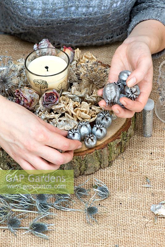 Creating a festive table decoration with garden flowers and seed heads. Place silvered nigella seed heads onto oak base.