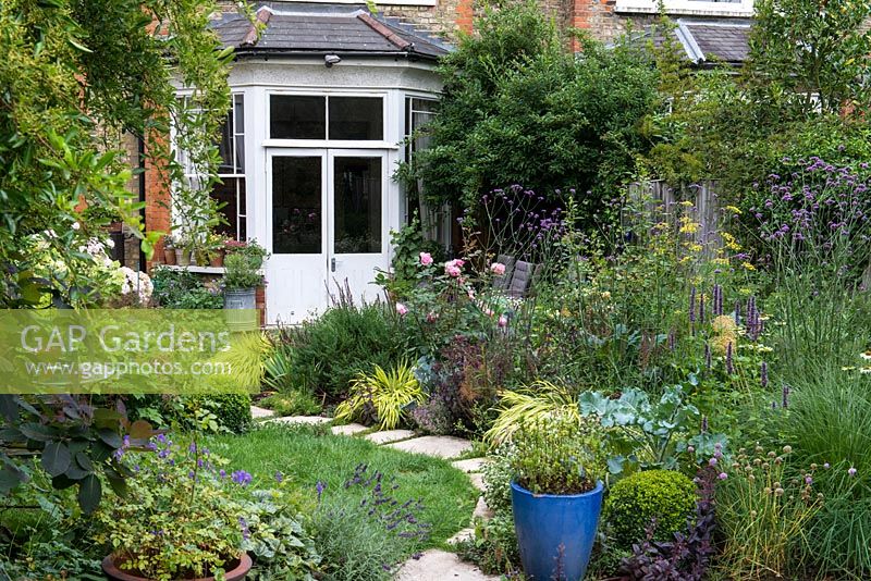 A small town garden with stone path, circular lawn and mixed borders planted with flowers, herbs and vegetables.