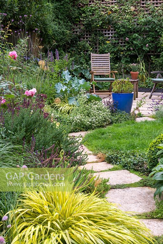 small walled town garden with patio seating area in front espaliered apple trees and gooseberry bushes. In the foreground, a harmoniously cloured mixed border with roses, salvia, rosemary, hackonechloa and Verbena bonariensis.