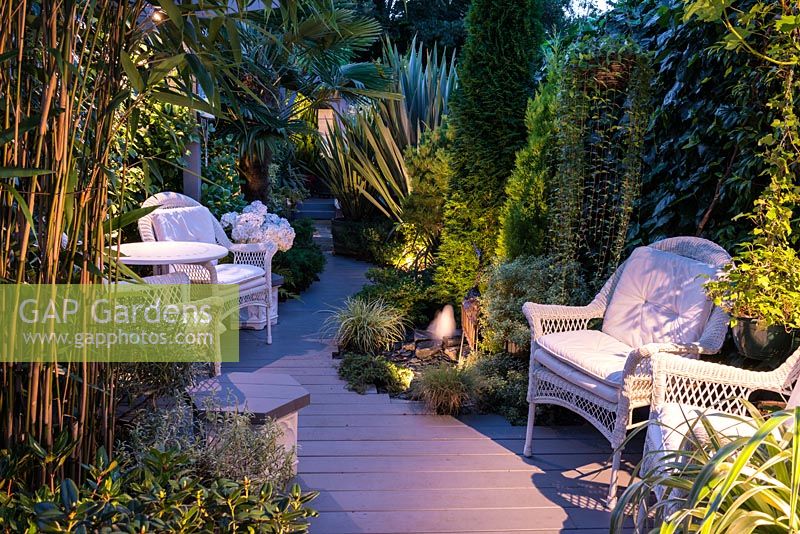 A small contemporary garden at night, with curved wooden path and seating area lit at night.