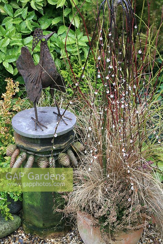 Muted brown and bronze tones for winter and spring. Rust coated cockerel sculpture made from scrap metal and showcased on a chimney pot topped with a circle of slate and decorated with a necklace of fir cones. Weeping pussy willow is underplanted with bud heathers and brown leaved sedge in a terracotta pot.