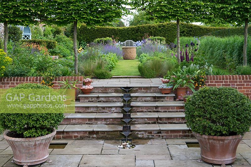 A contemporary, pumped water feature carries water down the steps to a small pebble pool. Above, pleached hornbeams frame views of perennial borders.