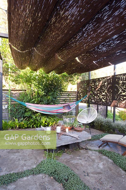 Seating area of garden showing timber table, decorative timber screen and hammock all under a retractable shade hut canopy
