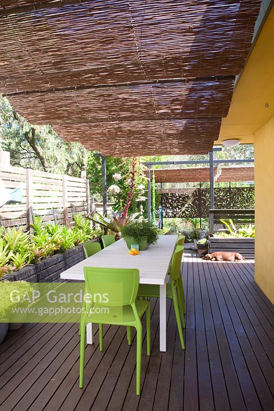 Timber deck with informal dining area and timber panelled screens. Table with potted rhipsalis, and raised bed with bromeliads seen