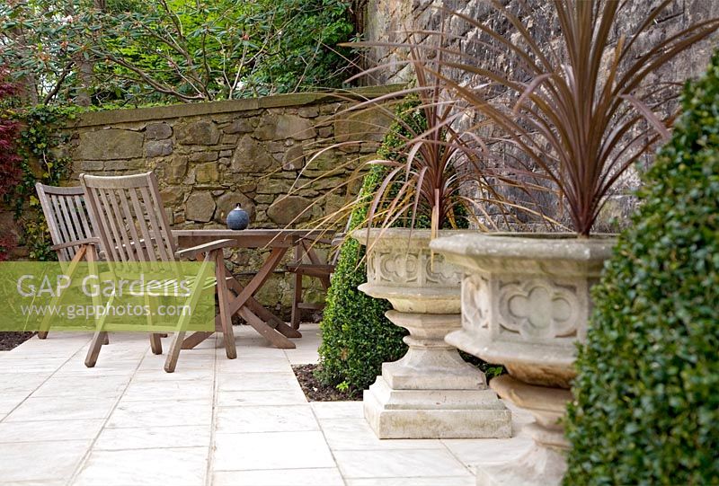 Paved seating area featuring cordylines in stone urn planters and box pyramids
