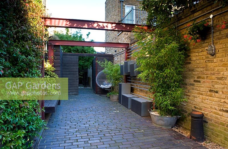 Alleyway beside house with wall lighting, bamboo in container galvanised bath, steel girder pergola and wooden fence with open gate at the end. Brick flooring. Metal storage boxes hung on a rack on the wall. 