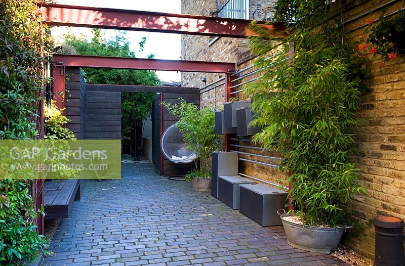 Alleyway beside house with wall lighting, bamboo in container galvanised bath, steel girder pergola and wooden fence with open gate at the end. Brick flooring. Metal storage boxes hung on a rack on the wall.

