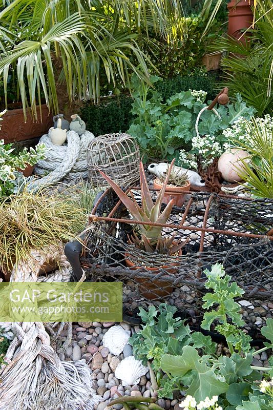 Seaside themed border with numerous ornamental set-dress items including crab pot, rope, anchor, buoys and planting including crambe maritima and chusan palm. 
