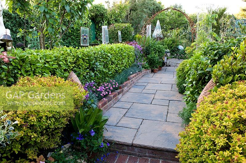 Paving path between borders planted with a tapestry of shrubs including golden spirea, bay, griselinia and edging of dianthus. Sculptural ornaments including stained glass panels. 