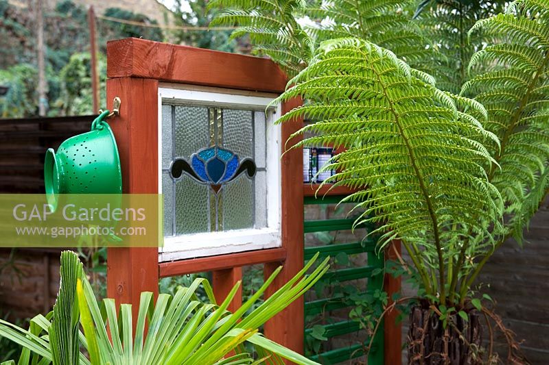 Dividing panel constructed from wood and reclaimed window frame with ornaments of found items including a kitchen colander with tree fern fronds and chusan palm