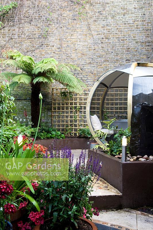 Low brown painted wall retaining raised beds, plants in containers including salvia and agapanthus, tree fern Dicksonia antarctica, high neighbouring boundary wall and seating inside a large round pod