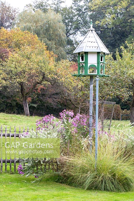 Wooden bird feeder standing over scene with perennials and grasses next to wooden picket fence. Pasture and apple trees in the background
