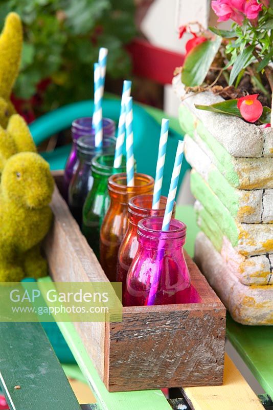 Detail of an inner city garden with artificial grass rabbit ornaments, retro cement pots, recycled timber box with coloured glass bottles with drinking straws and colourful outdoor table
