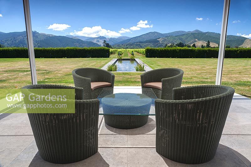 Rattan chairs and glass table with view to the mountains and vineyards beyond at Bhudevi Estate garden, Marlborough, New Zealand.