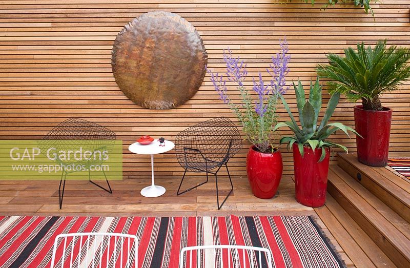 Brazilian hardwood decking, red containers with perovskia, agave and fern, table and chairs and copper beaten disc on wall. Ben De Lisi House and Garden, London

