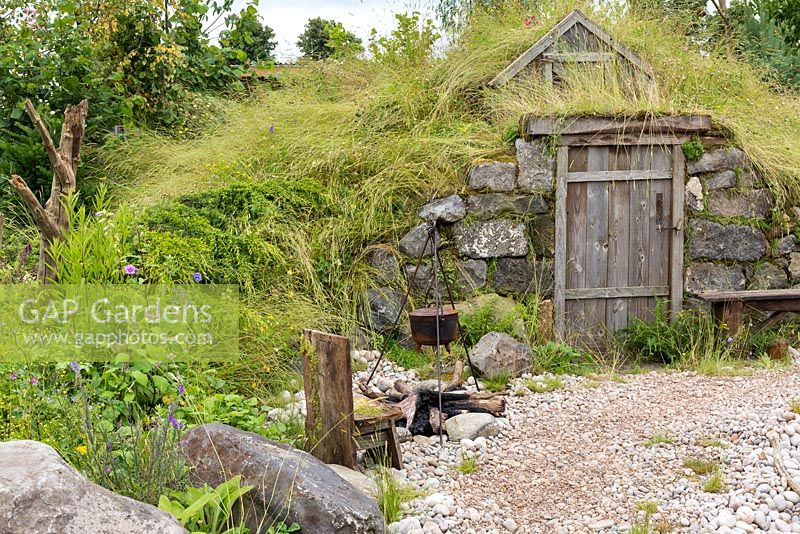 The Viking Cruises Scandinavian Garden, view of troglodytic hut built into hillside with beach pebbles in front and wild meadow with shaggy grasses above and to the sides. A rusted cooking pot on metal tripod sits over charred logs, on the shingle. RHS Hampton Court Palace Flower Show, 2016 