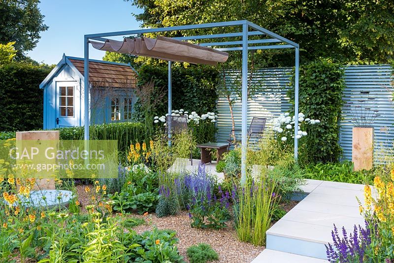 Retreat Garden, Steel pergola with canvas awning stands over metal table and chairs on white paved raised terrace area with gravel surrounding. Planting is direct into the gravel at the front, with drought tolerant plants including verbascum, echinacea, artemisia, geum, perovskia, stipa gigantea and salvias. RHS Hampton Court Flower Show in 2016