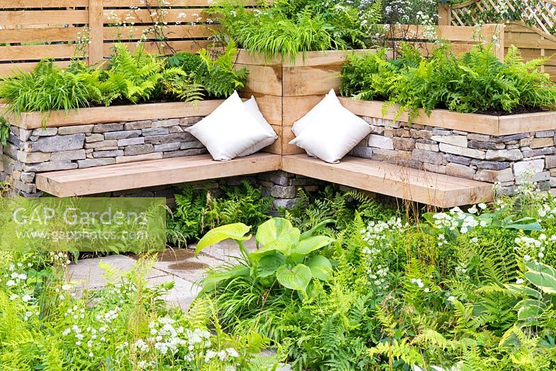 Inner City Grace, York stone and oak corner bench with white cushions provides a relaxing space in a city garden, surrounded by green planting including Hosta 'Patriot', Hosta 'Prince of Wales', Astrantia 'Great White' and Astrantia major 'Alba', grasses Deschampsia cespitosa and Hakonechloa macra, and ferns Dryopteris filix-mas, Dryopteris erythrosora 'Brilliance', and Dryopteris affinis 'Cristata The King'. RHS Hampton Court Flower Show in 2016