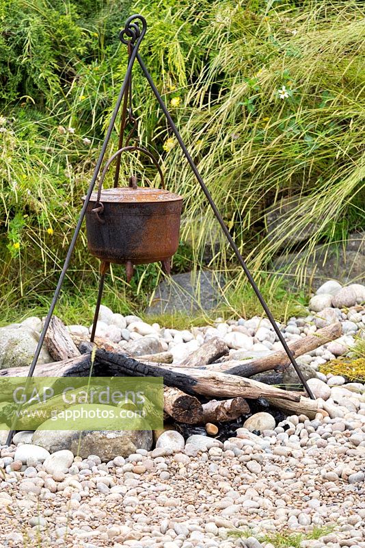 The Viking Cruises Scandinavian Garden, detail of rusted cooking pot on metal tripod over charred logs, on pebble and shingle , with wildflower meadow behind. RHS Hampton Court Flower Show 2016