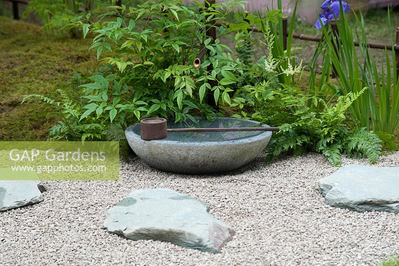 Detail of a 'Japanese Summer Garden' designed by Saori Imoto showing the gravel path set with granite stepping stones, on which sits a stone bowl for water with a bamboo spout and wooden ladle. Planting includes moss, nandina, ferns and iris. A low bamboo fence borders the garden on one side. Hampton Court Flower Show, July 2016.