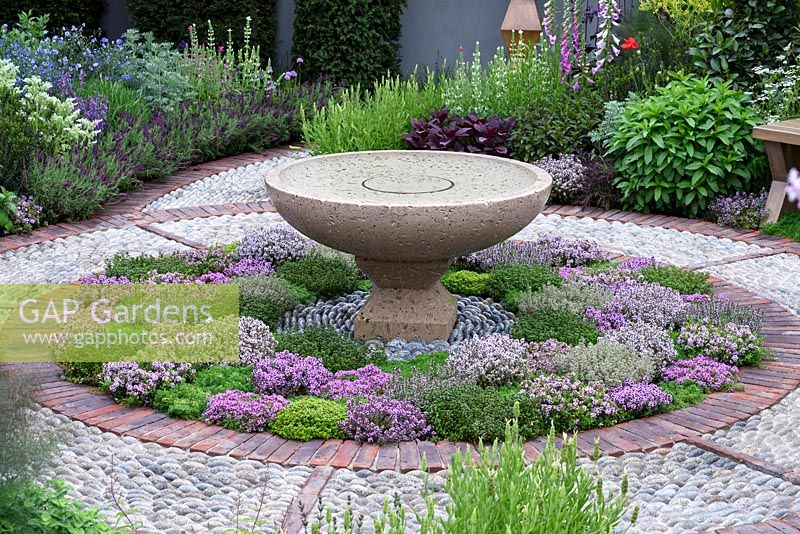 The St John's Hospice Garden, The Modern Apothecary. Extensive planting of herbs around water basin in circular bed with cobble stone surround. RHS Chelsea Flower Show 2016. Designer: Jekka McVicar, Sponsor: St John's Hospice